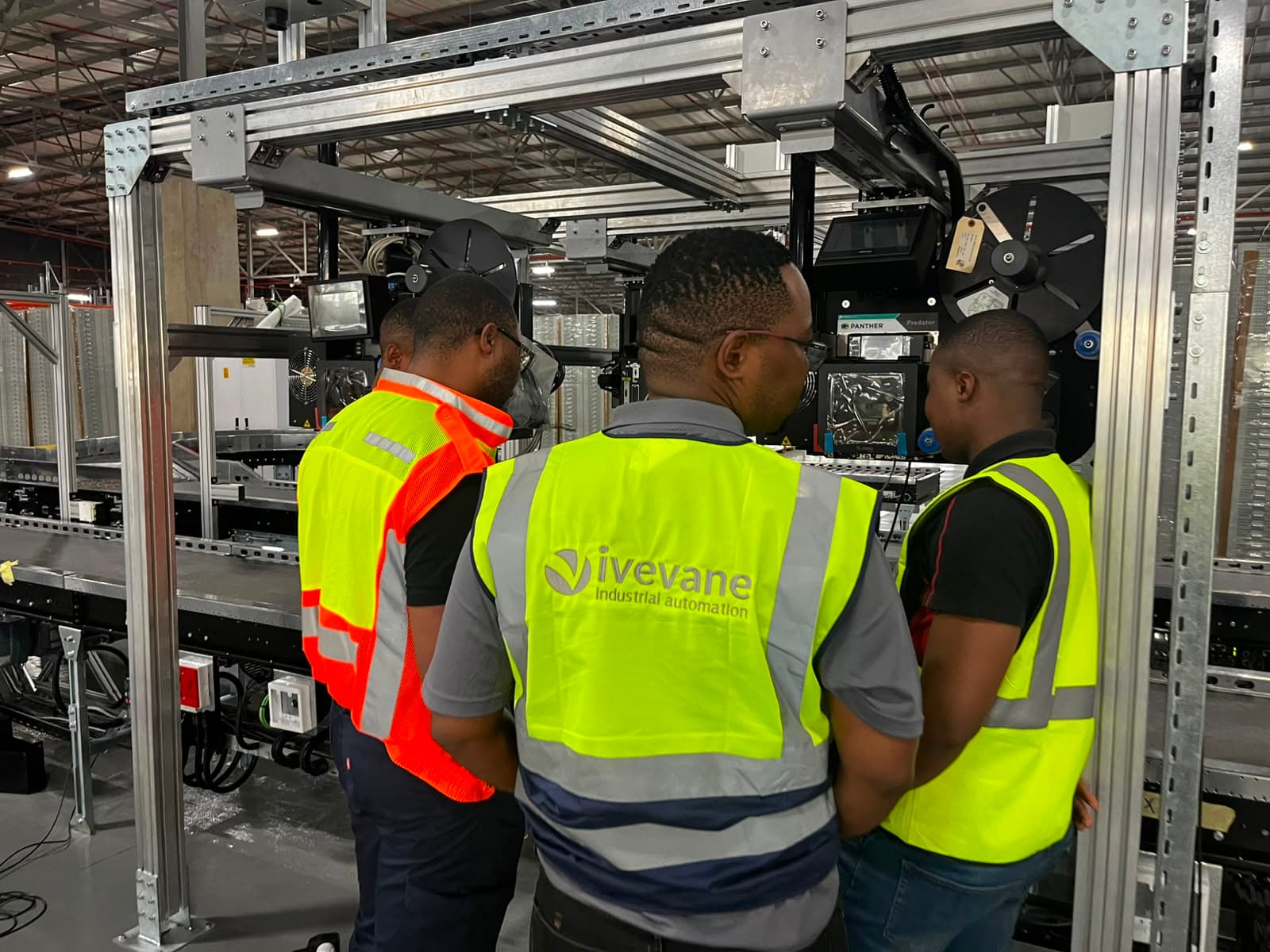 Three Ivevane engineers in branded high-visibility vests reviewing a Panther print-and-apply labelling line during commissioning inside a distribution facility.