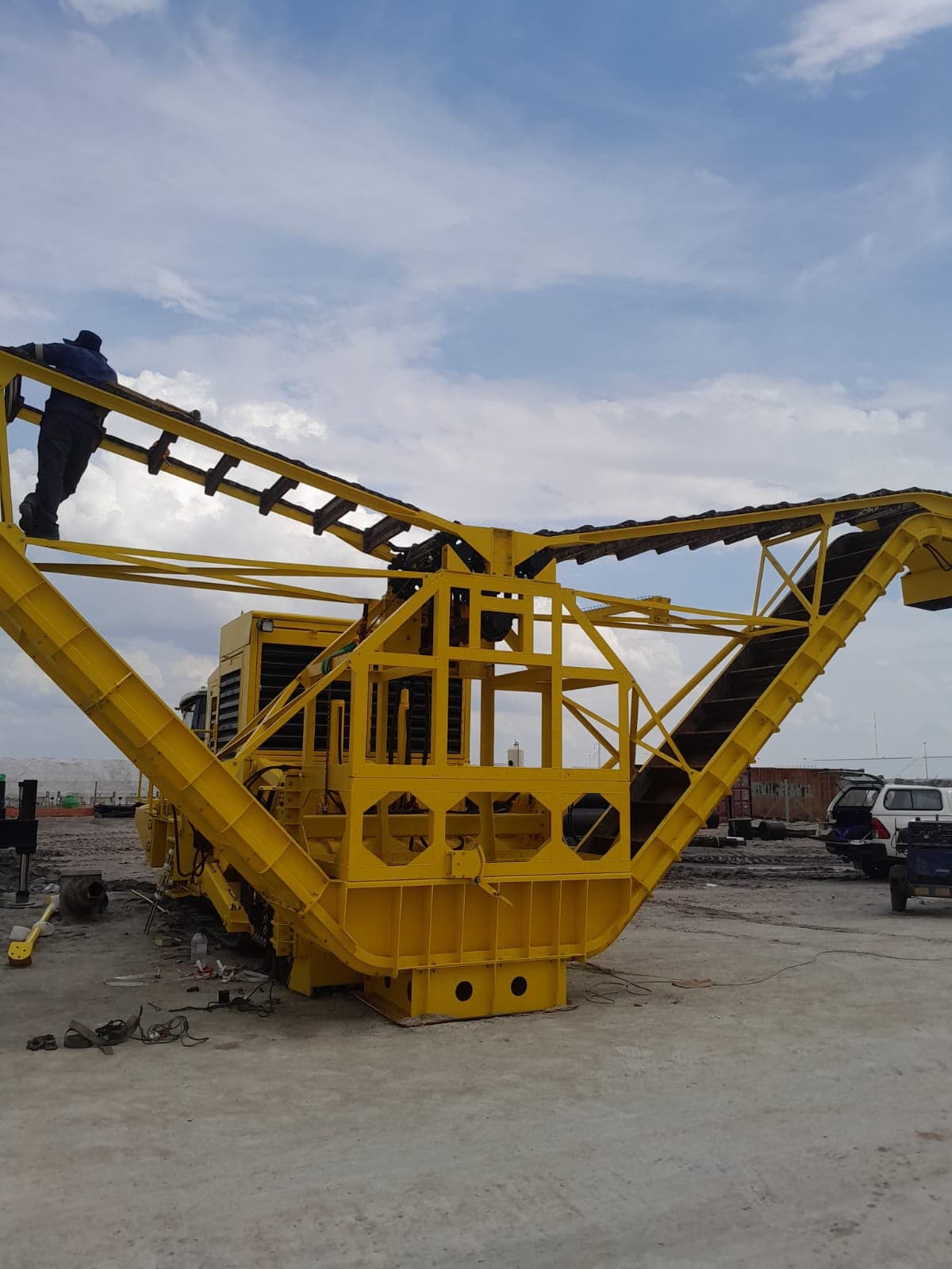 A large yellow salt harvester under assembly on a mine pan, an engineer standing on its conveyor boom giving scale against an open sky.