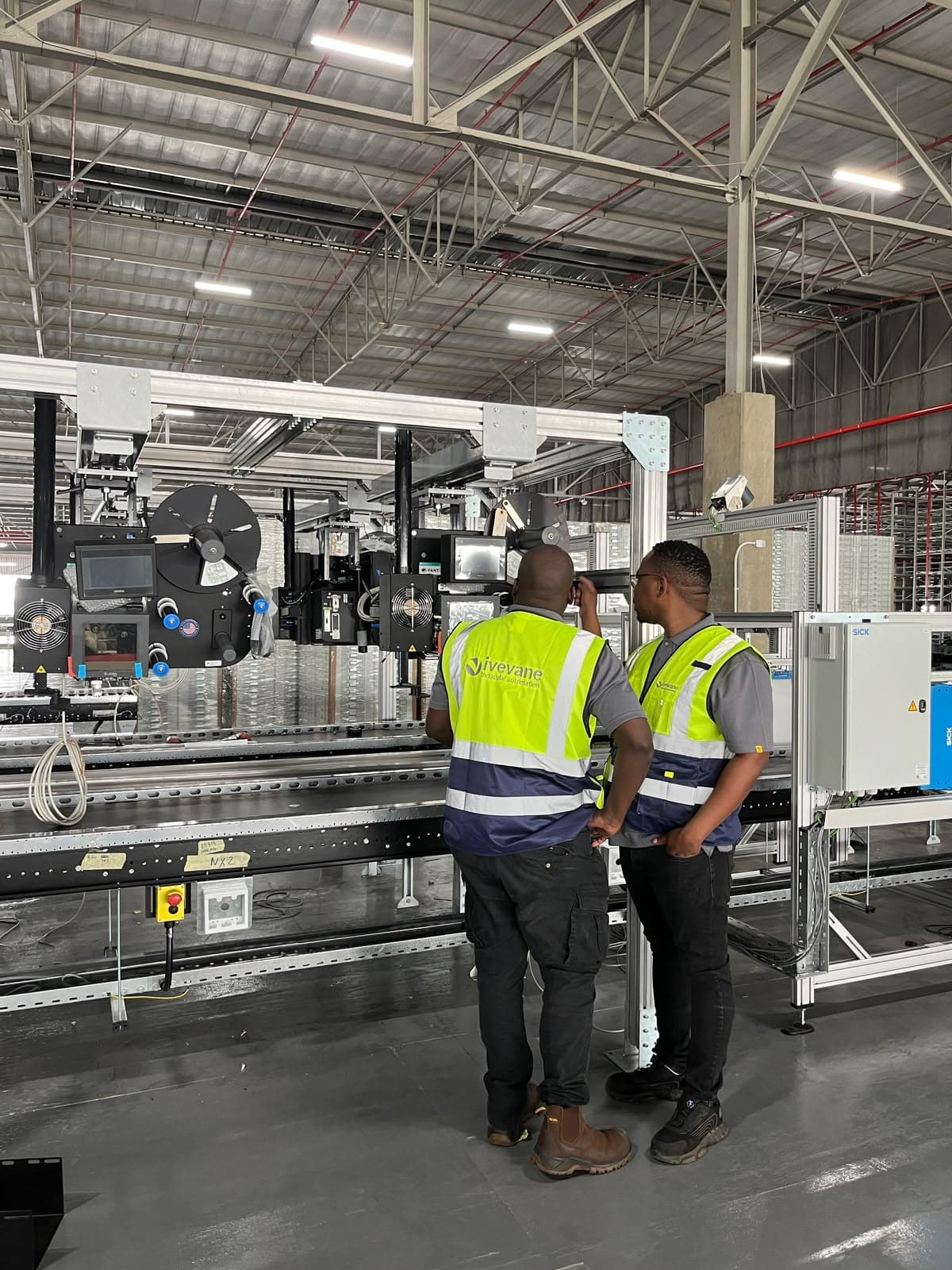 Two Ivevane engineers in branded high-visibility vests commissioning an overhead print-and-apply labelling head above a conveyor line.