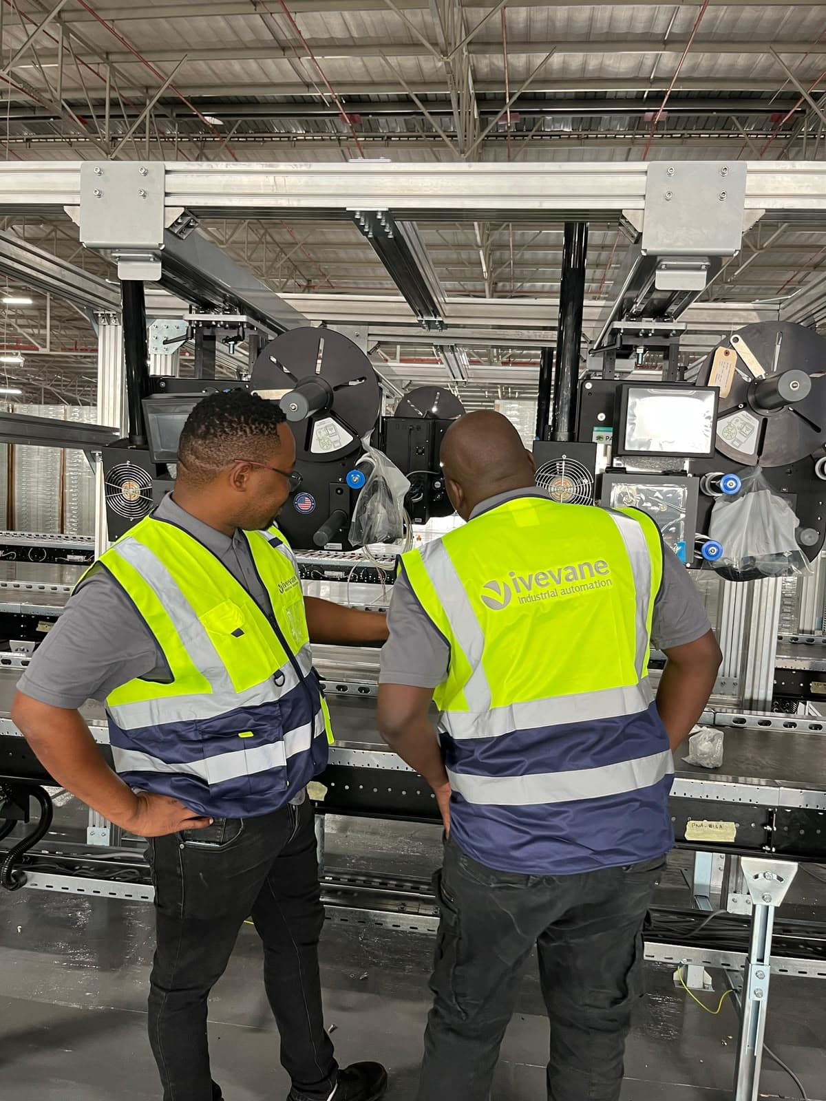 Two Ivevane engineers in branded high-visibility vests working together at a labelling line, the company logo clearly visible on the vest.
